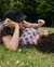 A couple lying on the grass, facing each other. The person on the left, wearing a black hat, is resting their head on their hand while looking at their partner, who is lying down beside them. They are surrounded by a lush green park with trees in the background